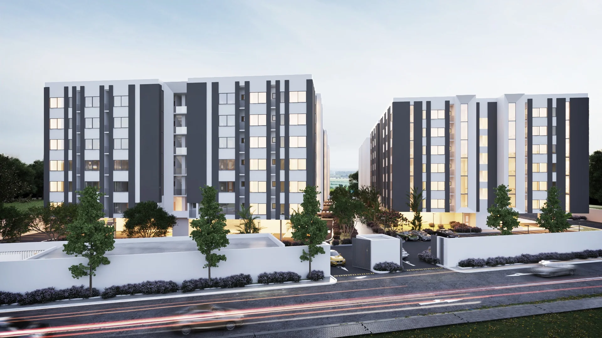 Two contemporary apartment blocks with dark vertical fins and warm-lit windows at dusk, beside a landscaped road